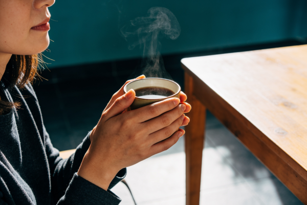 A woman cradling a warm, steaming cup in both hands while sitting by a wooden table